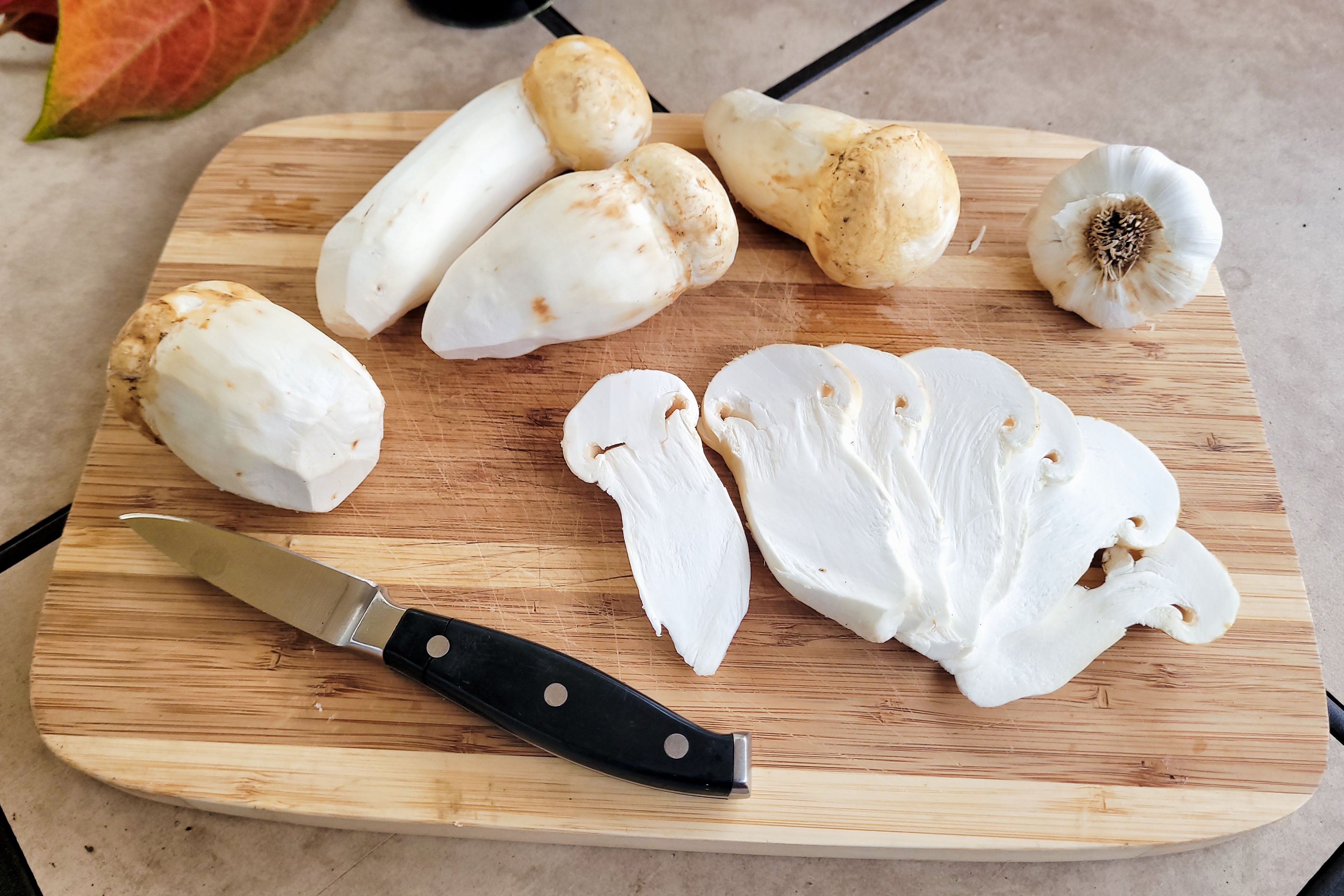 Fresh wild foraged matsutake mushrooms displayed on a wooden cutting board with a knife and a small pile of shredded matsutake flesh in the background.